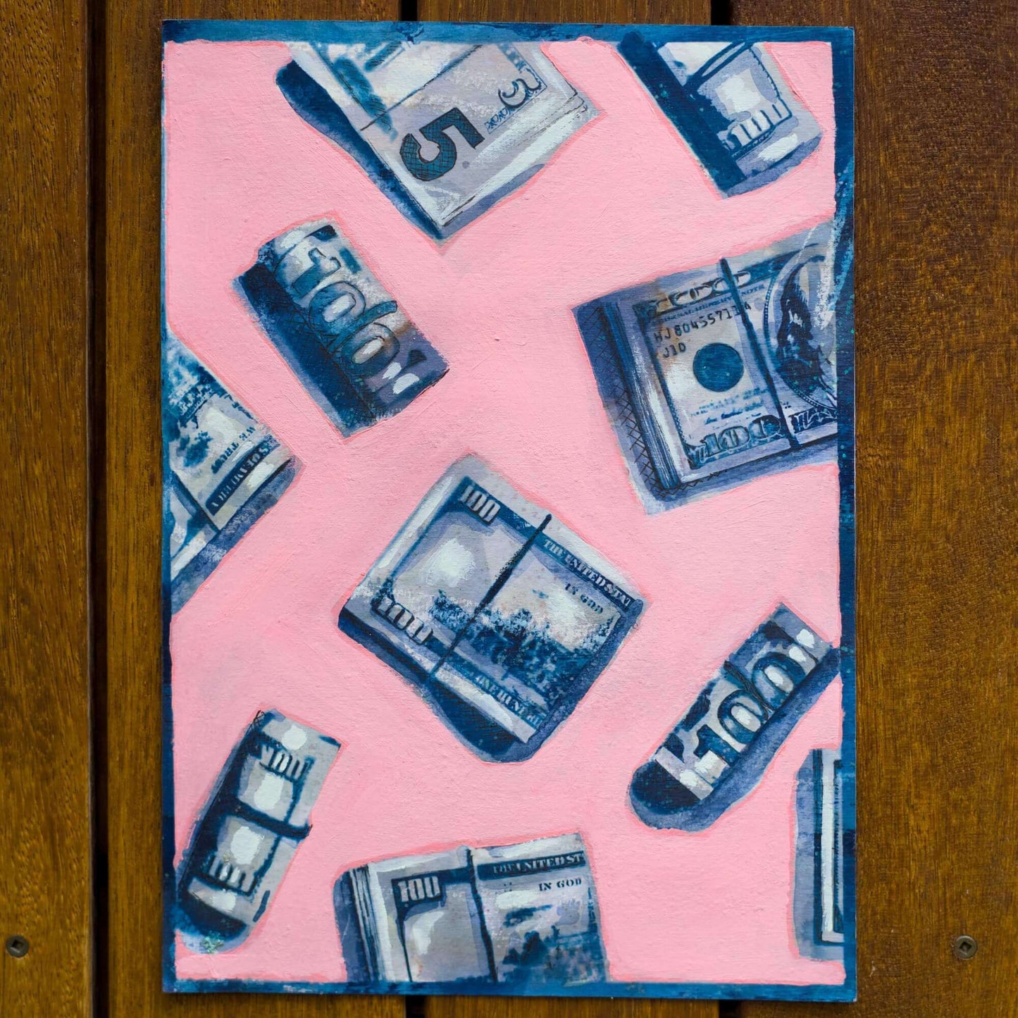 A framed artwork featuring a repeating pattern of blue and white dollar bills on a pink background