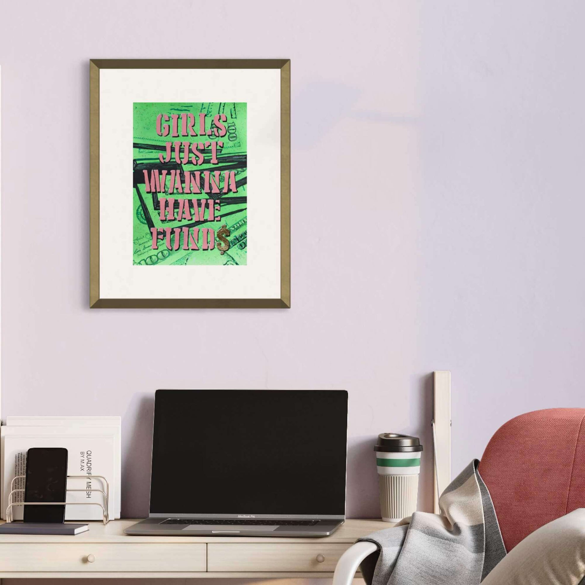 A framed artwork with a neon green background and the text 'Girls just wanna have funds' displayed on a wall above a desk with a laptop and other items.