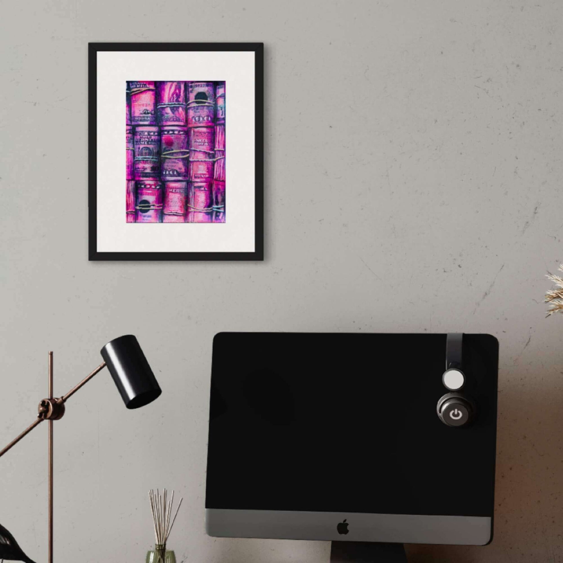 A framed abstract painting featuring pink and purple rolls of money, displayed on a white wall above a black desk with a computer monitor and decorative lamp.
